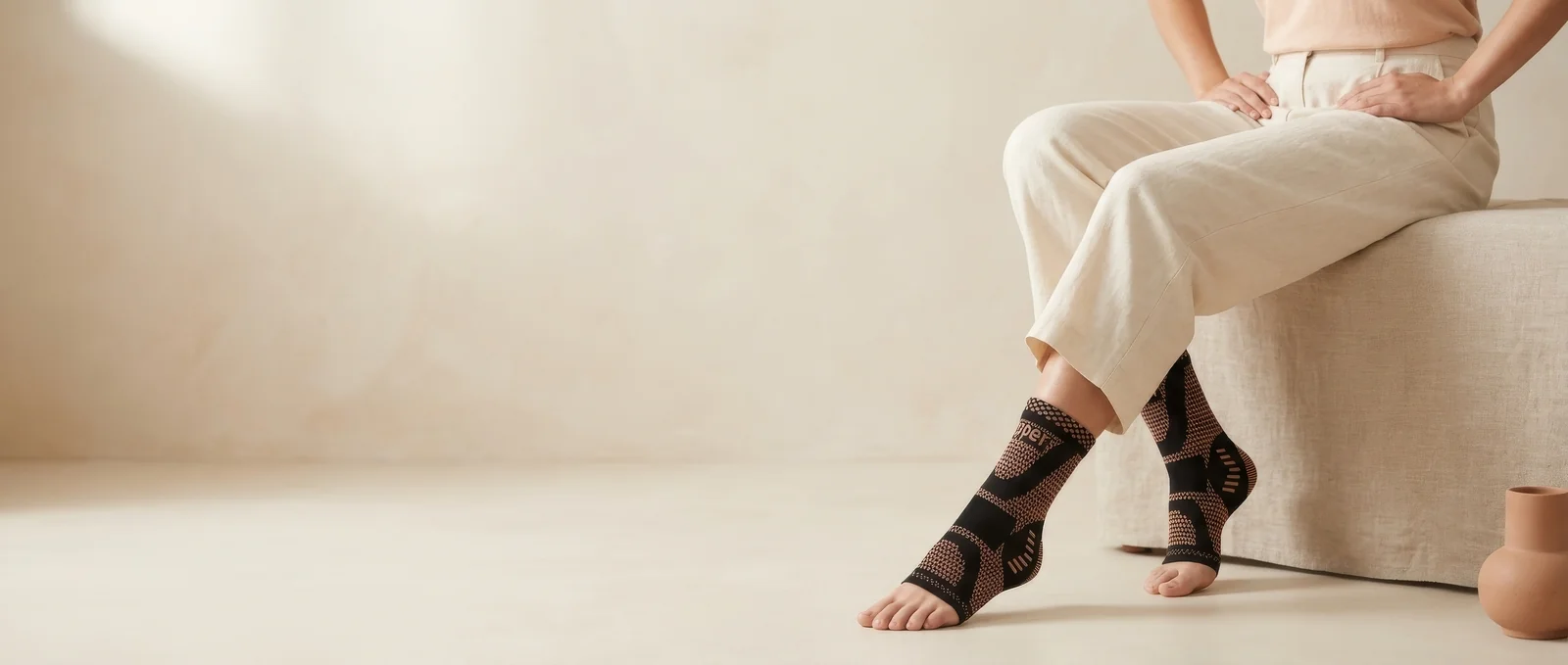 FreeStep™ compression socks worn with cream linen trousers, seated on a linen-covered bench in a warm cream interior, premium editorial ultrawide crop